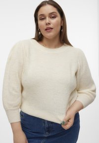 Maglione lavorato a maglia crema con scollo rotondo, maniche voluminose e orlo a coste. Abbinato a una gonna di jeans blu, che mette in risalto la morbidezza della texture.