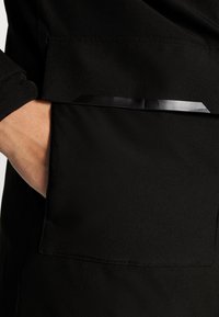 Tessuto nero con una trama liscia presenta una tasca e una striscia di accento nera lucida. La vista ravvicinata mette in evidenza i dettagli della cucitura.