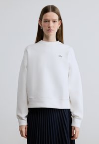 Sweat-shirt en coton blanc avec un col rond, des poignets côtelés et un petit logo brodé. Associé à une jupe plissée foncée.