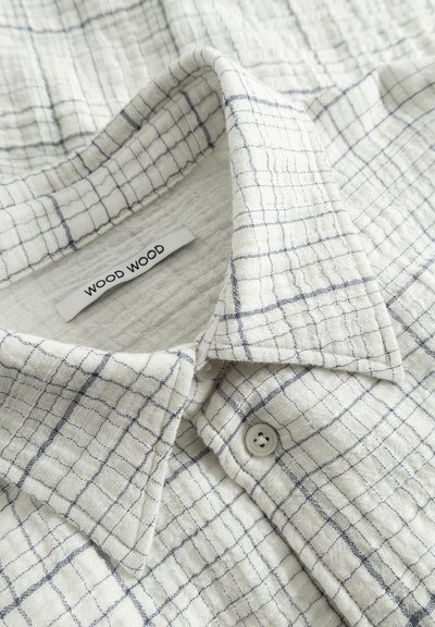 Chemise à carreaux blanche et bleue à boutons, fabriquée en tissu texturé, avec un col rabattu et deux boutons visibles sur le devant.