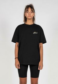 Black cotton t-shirt with a round neck and short sleeves, featuring a logo in gold. Worn with black biker shorts, both in a casual fit.
