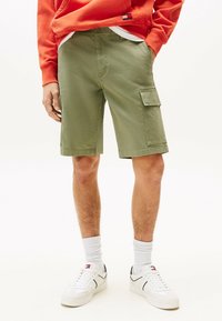 Pantalones cortos cargo verdes con bolsillos laterales, hechos de algodón. Combinados con zapatillas blancas y calcetines acanalados. Modelo vistiendo una sudadera roja.