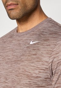 Camiseta atlética marrón jaspeada de manga corta, con cuello redondo y un logotipo blanco de Nike en la parte izquierda del pecho. Textura suave.