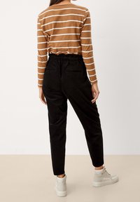 Kvinna som bär en brun och vit randig långärmad topp, svarta högmidjade byxor och vita högskurna sneakers, stående med ryggen mot kameran.