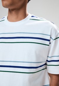 T-shirt bianca a maniche corte con strisce orizzontali blu, verdi e azzurro chiaro. Girocollo e tessuto morbido in cotone.