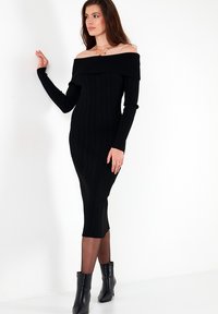 Musthave mustribaline musta midi kleit, mis jääb õlgadest vabaks, pikaaegsete varrukatega, keha ligi liibuva siluetiga ja külgmise lõhikuga, kombineeritud mustade pahkluu saapadega.