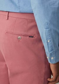 Himmelblaues Button-up-Hemd kombiniert mit rosa Baumwollhosen, die über eine Gesäßtasche mit einem Knopf und einem sichtbaren Etikett verfügen.