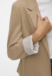 Beige op maat gemaakte blazer met een soepele textuur, voorzien van witte en gouden gestreepte opgerolde mouwen. Daaronder is een simpele witte top gedeeltelijk zichtbaar.