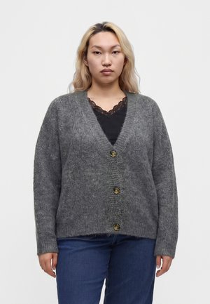 Junge Frau mit blondem Haar trägt eine graue, geknöpfte Strickjacke über einem schwarzen Spitzen-Top und blauen Jeans, stehend vor einem einfachen weißen Hintergrund.