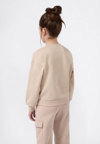 Femme avec des cheveux bruns en chignon, portant un sweat-shirt beige et un pantalon cargo, vue de dos sur un fond blanc uni.