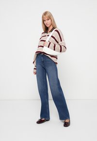 Beanie-gestreepte trui in beige en bordeaux, gecombineerd met uitlopende blauwe jeans. Het model draagt ook een witte tas en heeft donkere schoenen aan.