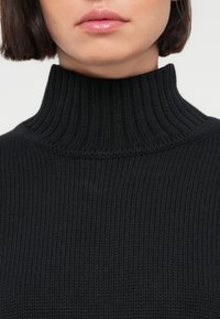 Gros plan d'une personne portant un pull à col roulé côtelé noir, montrant les lèvres ainsi qu'une partie du cou et du menton.