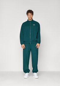 Ensemble de survêtement teal avec veste à zip et poignets élastiques, pantalons assortis avec logo sur la jambe, fabriqué dans un matériau doux, associé à des baskets blanches.