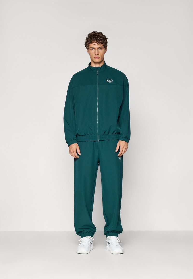 Ensemble de survêtement teal avec veste à zip et poignets élastiques, pantalons assortis avec logo sur la jambe, fabriqué dans un matériau doux, associé à des baskets blanches.