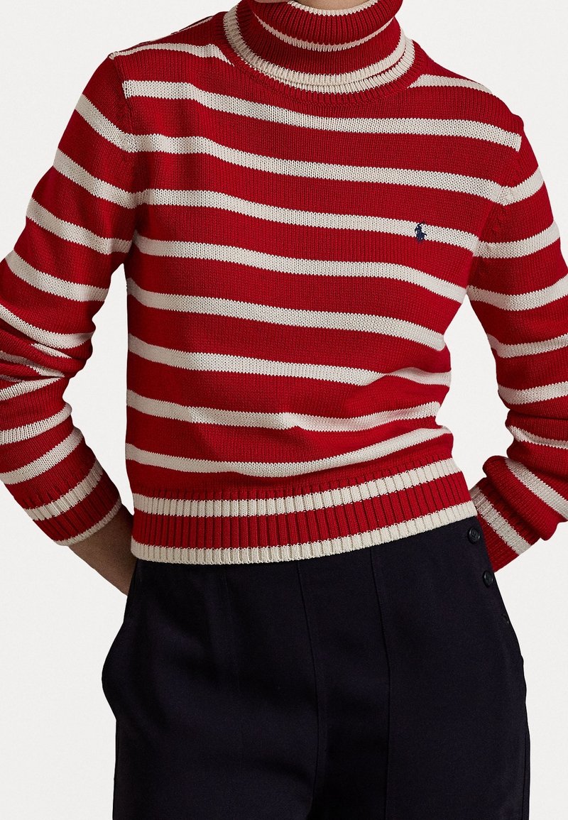 Rot-weiß gestreifter Strickpullover mit hohem Kragen und gerippten Bündchen; kleines Logo auf der Brust; kombiniert mit dunklen Hosen mit Knopfakzenten.