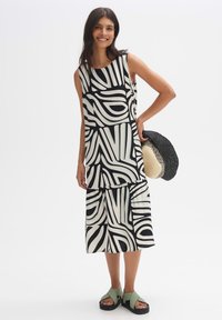 Sleeveless dress in a black and white abstract pattern, with a tiered design and flowy fabric. Paired with a round straw hat and sandals.