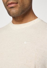 Camiseta de manga corta beige hecha de tejido texturizado, que presenta un pequeño logotipo reflectante de Nike en el área del lado izquierdo del pecho.
