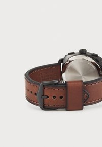 Bracelet de montre en cuir marron avec bouclerie en métal noir. Présente des détails de couture et plusieurs trous pour l'ajustement. Dos de montre en acier inoxydable visible.