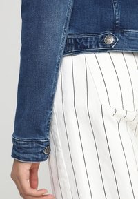 Tummansininen farkkutakki, jossa on ommellut saumat ja metalliset napit; yhdistetty valkoisiin pystysuoriin raidallisiin housuihin. Pehmeä pinta.