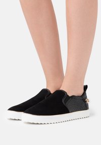 Czarne wsuwane buty z zamszu z teksturowanym czarnym akcentem, białą podeszwą i niewielkim złotym elementem ozdobnym. Lekko podwyższony obcas.