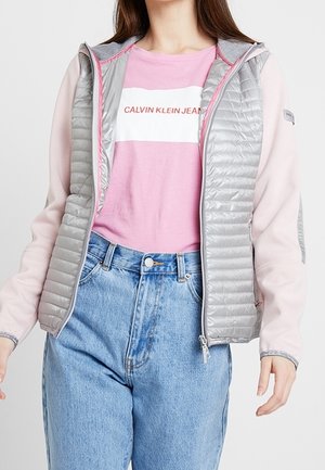 Femme portant un gilet matelassé gris clair sur un t-shirt Calvin Klein Jeans rose et blanc avec un jean taille haute bleu clair.