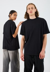 Deux individus portent des t-shirts noirs surdimensionnés. L'arrière-plan est uni. Les t-shirts ont une coupe décontractée et un col rond. La femme a les cheveux relevés en chignon.