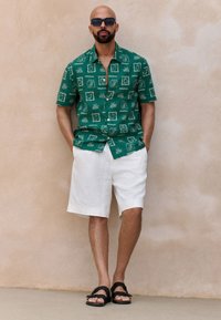 Green short-sleeve shirt with white patterned accents, paired with light beige shorts and black sandals. Smooth fabric texture, relaxed fit.