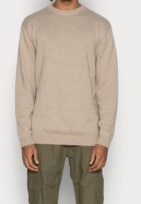 Maglione beige a maglia con collo rotondo a coste, maniche lunghe e un motivo di tessuto testurizzato, abbinato a pantaloni cargo verde oliva.