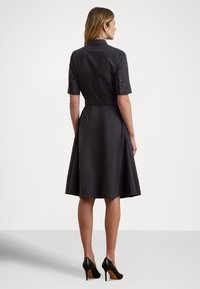 Vestido preto de manga curta com cintura ajustada, saia evasê e detalhes de punho com botões. Usado com sapatos de salto alto pretos.