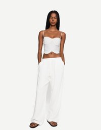 Une personne avec de longues tresses se tient avec confiance, portant un crop top blanc et un pantalon blanc ample, devant un fond uni.