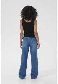 Eine Person mit lockigem Haar steht mit dem Rücken zur Kamera, trägt ein schwarzes Tanktop und weite blaue Jeans vor einem schlichten Hintergrund.
