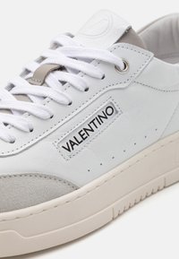 Hvide lædersneakers med grå ruskindsdetaljer, udstyret med flade hvide snørebånd, et fremtrædende logo-label og en struktureret gummisål.