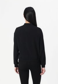 Donna con lunghi capelli neri che indossa un top nero a maniche lunghe e pantaloni neri, in piedi di fronte a uno sfondo bianco.