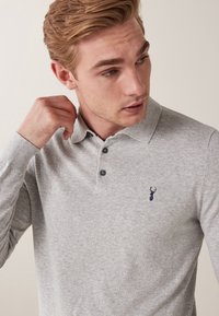 Harmaa pitkähihaisen poolopaidan paita, jossa on kaulus, kolme tummaa nappia ja pieni laivastonsininen peuran logo rinnassa. Pehmeä puuvillakangas.