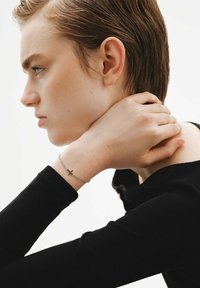 Pulsera de plata con un pequeño colgante de cruz, llevada en una muñeca pálida, con una camisa de manga larga negra visible de perfil.