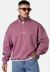 Sweat-shirt rose à demi-zip, de coupe décontractée, avec des poignets côtelés et un logo brodé sur la poitrine. Le tissu semble doux et texturé.