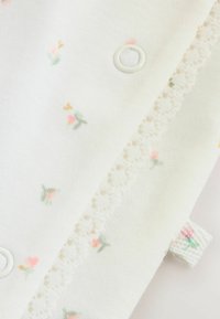 Biały materiał z małymi kwiatowymi wzorami w kolorach różowym i zielonym, z delikatną koronkową lamówką oraz okrągłym zatrzaskiem. Miękka tekstura.