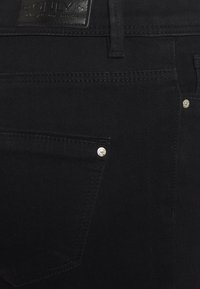 Calças de ganga pretas com um acabamento texturizado, costuras visíveis, um pequeno rebite prateado e uma etiqueta de couro na cintura.