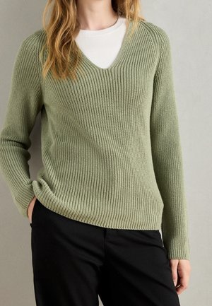 Frau trägt hellgrünen gerippten V-Ausschnitt-Pullover über weißem Top und schwarzen Hosen, mit einer Hand in der Tasche, vor einfarbig grauem Hintergrund.