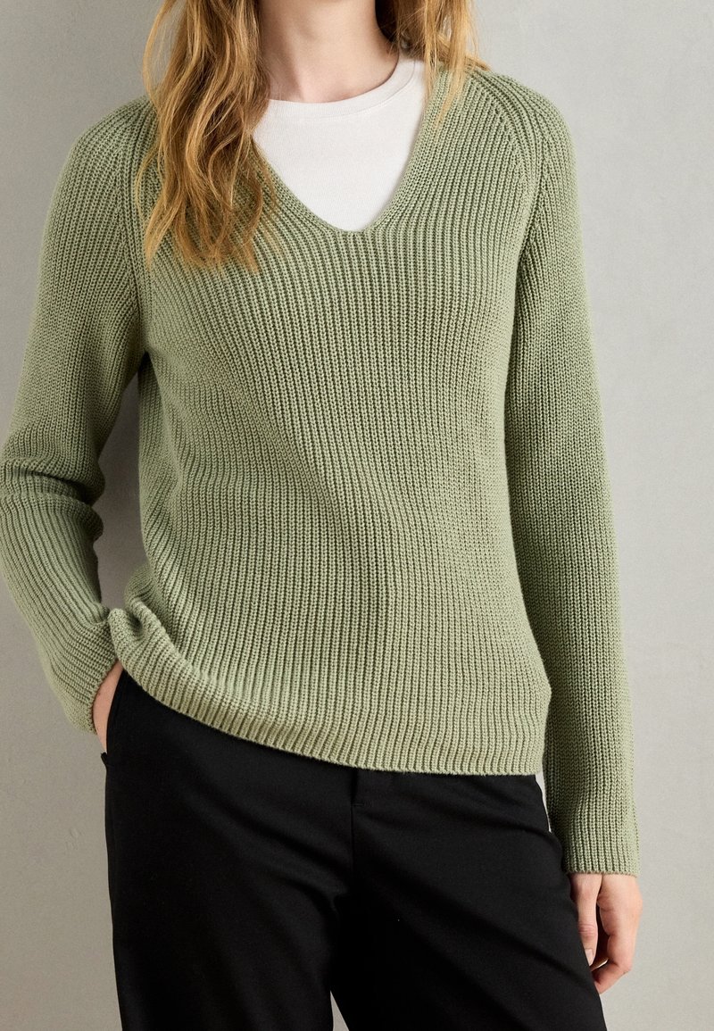 Femme portant un pull à col en V en maille côtelée vert clair sur un top blanc et un pantalon noir, avec une main dans la poche, devant un fond gris uni.