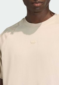 Beżowy bawełniany t-shirt z okrągłym dekoltem i krótkimi raglanowymi rękawami. Na piersi znajduje się subtelne, haftowane logo. Gładka faktura.