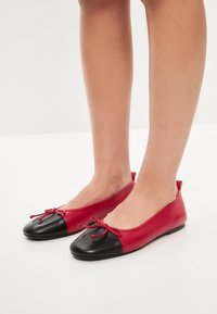 Ballerine in pelle rossa e nera con punta tonda, dettaglio del fiocco annodato e una superficie liscia con una punta a contrasto.