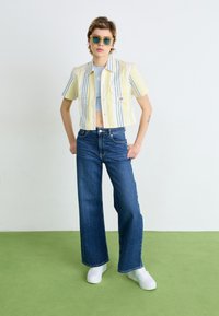 Cropped yellow and blue striped shirt, light blue underlayer, blue wide-leg jeans, white sneakers, green floor background.
