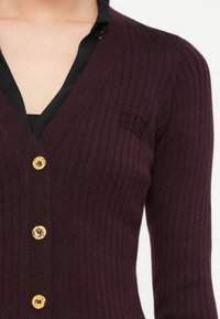 Close-up of a person wearing a ribbed deep purple cardigan with gold buttons over a black top with a V-neckline.