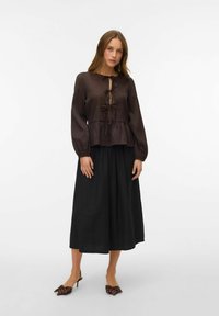 Tamno smeđa bluzica s peplum krojem, okruglim izrezom i detaljem mašne sprijeda, uparena s crnom, lepršavom suknjom srednje dužine i cipelama s oštrim vrhom.