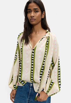 Kobieta nosząca luźną bluzkę z długim rękawem z pionowymi zielonymi, czarnymi i różowymi paskami, zestawioną z niebieskimi dżinsami.