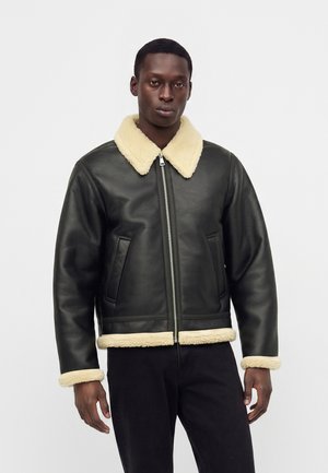 Jeune homme portant une veste en cuir noire avec col, poignets et ourlet en peau lainée beige, debout devant un fond clair uni.