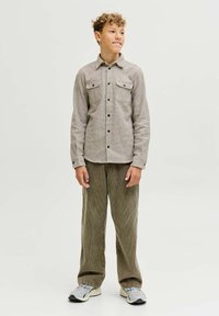 Camicia intrecciata color marrone chiaro con due tasche sul petto, apertura frontale con bottoni e colletto, abbinata a pantaloni larghi in corduroy verde e sneakers grigie.