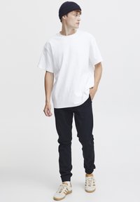White cotton t-shirt with a relaxed fit, paired with black tapered pants and beige sneakers featuring brown accents. Model wears a navy beanie.