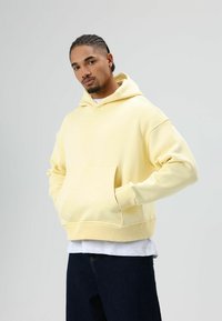 Gele capuchontrui gemaakt van zachte stof, met een grote frontzak, een losse pasvorm en geribbelde manchetten. Draagt over een wit shirt.